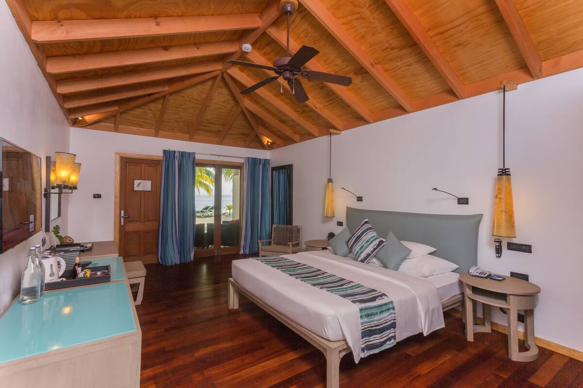 A spacious bedroom with a wooden ceiling, a bed with a striped blanket, bedside tables, and a desk with a lamp.