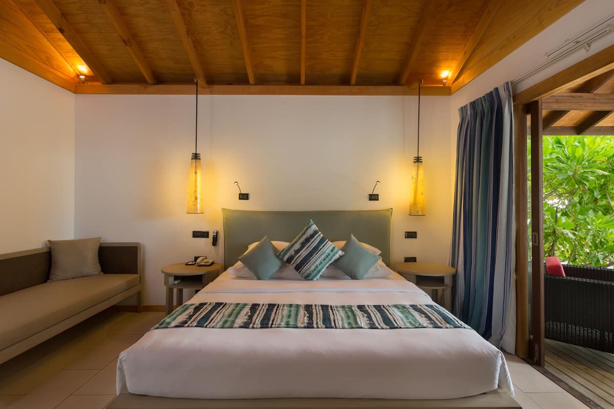 A bedroom with a large bed, two bedside tables, a sofa, and a view of greenery through a sliding door.