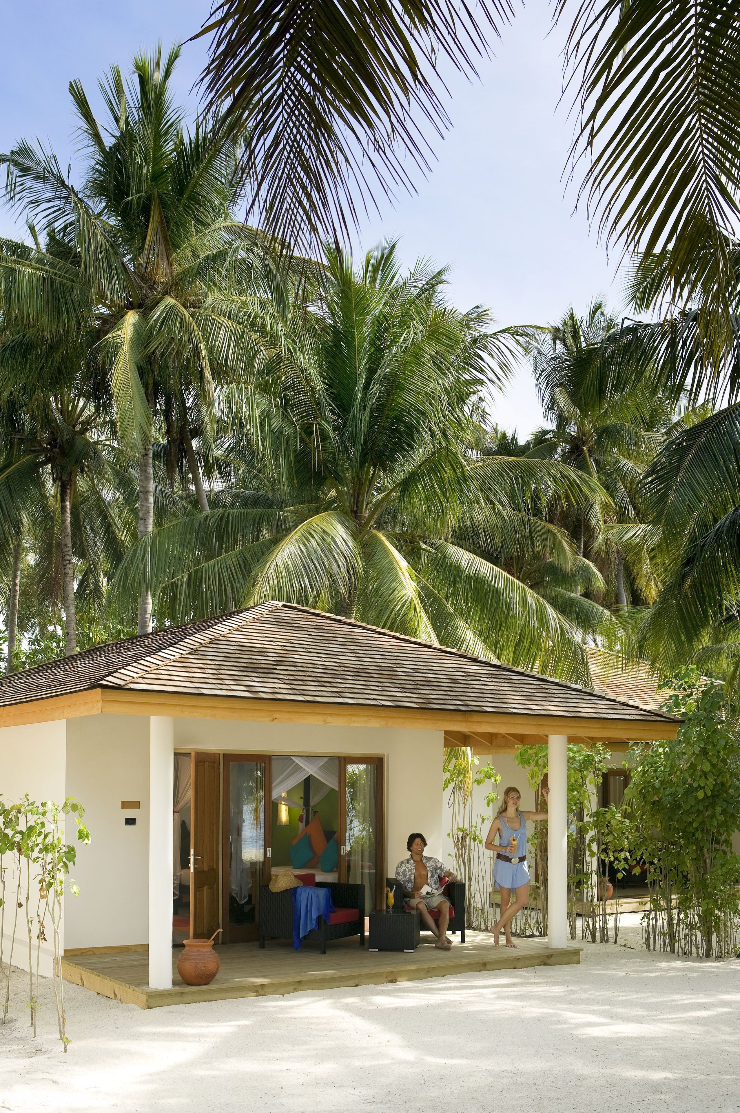 A small, single-story building with a tiled roof and a covered porch. There are palm trees in the background, and a few people are visible inside and outside the building.