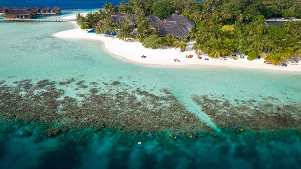 An aerial view of a tropical resort with overwater bungalows, a white sandy beach, and clear turquoise waters.