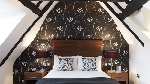 A bedroom with a sloped ceiling, a wooden headboard, and a patterned wallpaper. There are two bedside tables with lamps, a bed with a black and white duvet, and a carpeted floor. - The Boleyn Hotel (Staines)