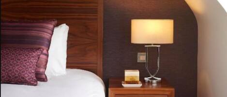 A bedroom with a wooden headboard, a bed with a white and purple striped bedspread, a nightstand with a lamp, and a small table with a book.