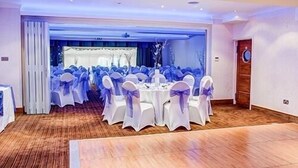 A banquet hall with round tables and chairs, white tablecloths, and blue sashes. - The Boleyn Hotel (Staines)