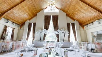 A dining area with wooden ceiling beams, large windows with curtains, and a chandelier. Tables are set with white tablecloths and glassware.