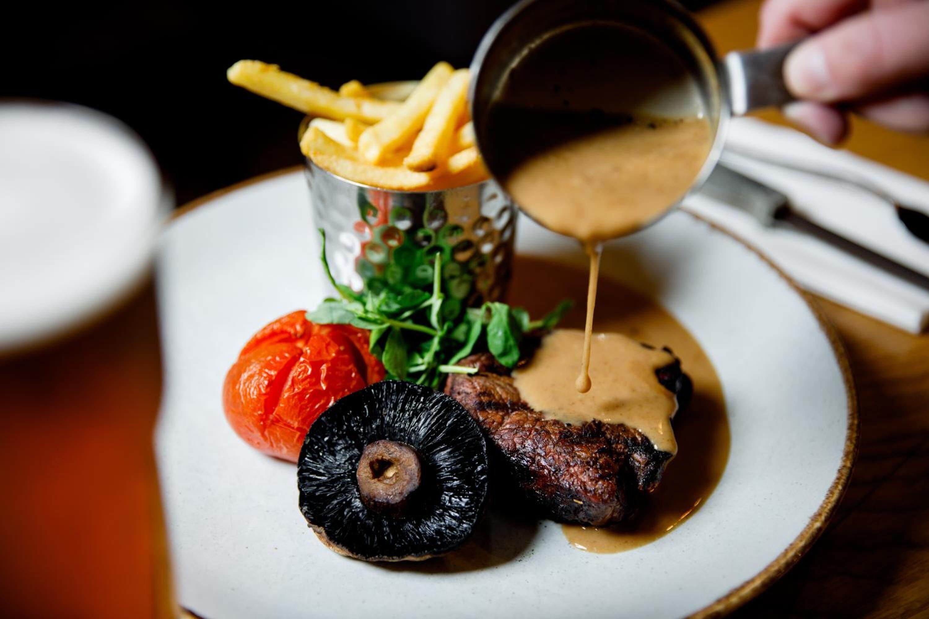 Ein Teller mit einem Steak, Pommes, einer Tomate und einem Pilz, während Sauce über das Steak gegossen wird.