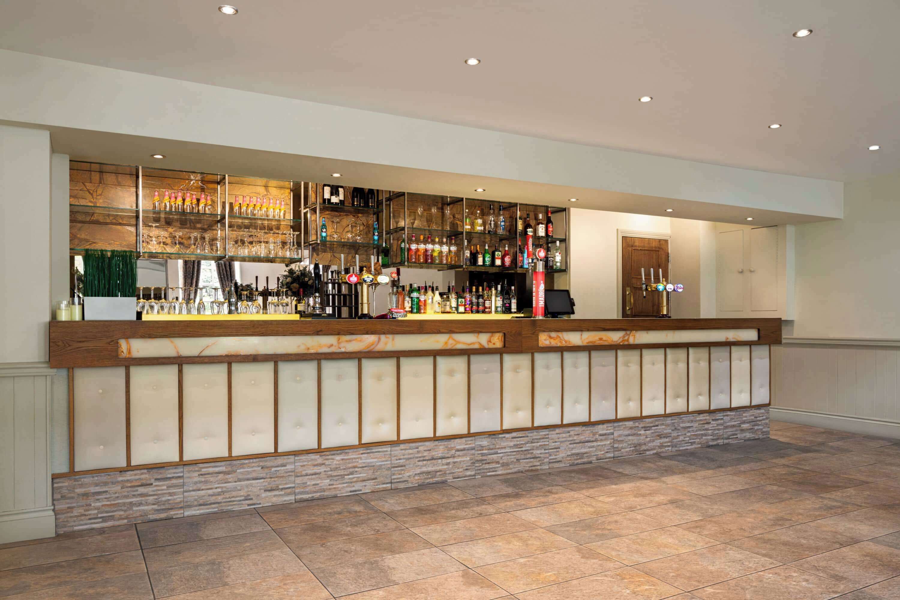 A bar area with a long counter, shelves with bottles and glasses, and a door to the right.