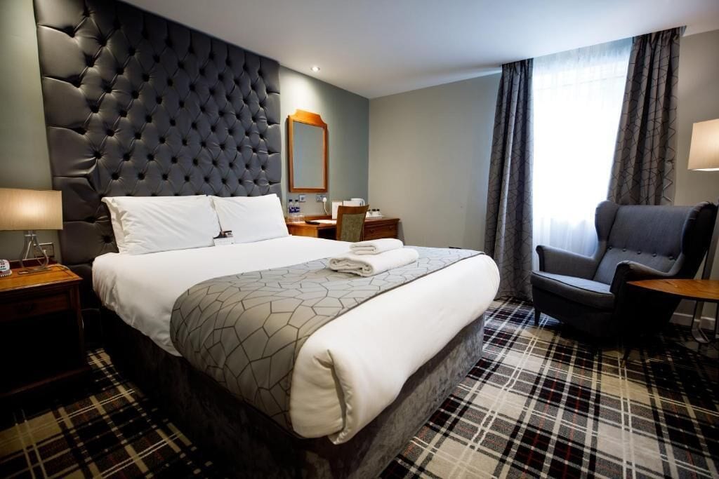 Ein Hotelzimmer mit einem großen Bett, einem gepolsterten Kopfteil, einem Sessel, einem Schreibtisch und einem Fenster mit Vorhängen.