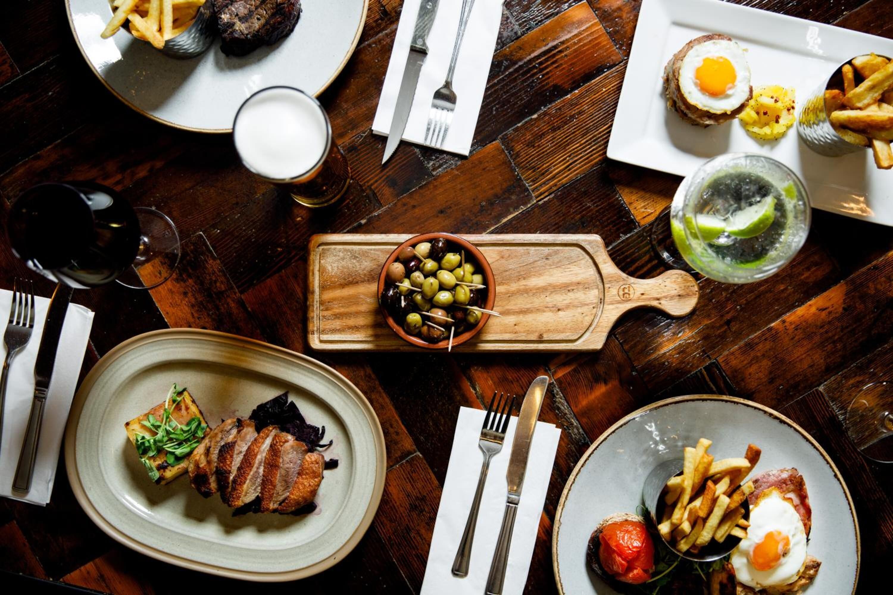 Une table à manger avec différents plats, notamment un steak, des frites et un bol d’olives.