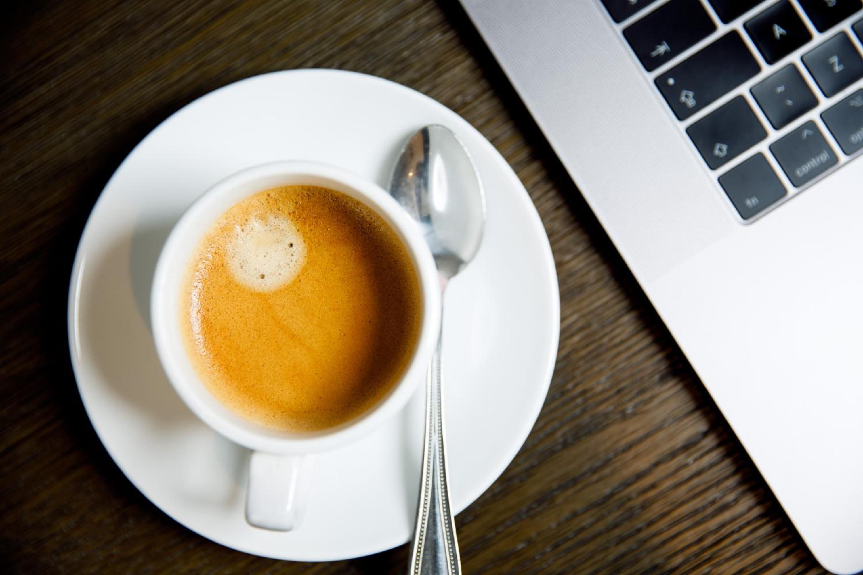 Una taza de café sobre un platillo con una cuchara, colocada sobre una mesa de madera junto a una computadora portátil.
