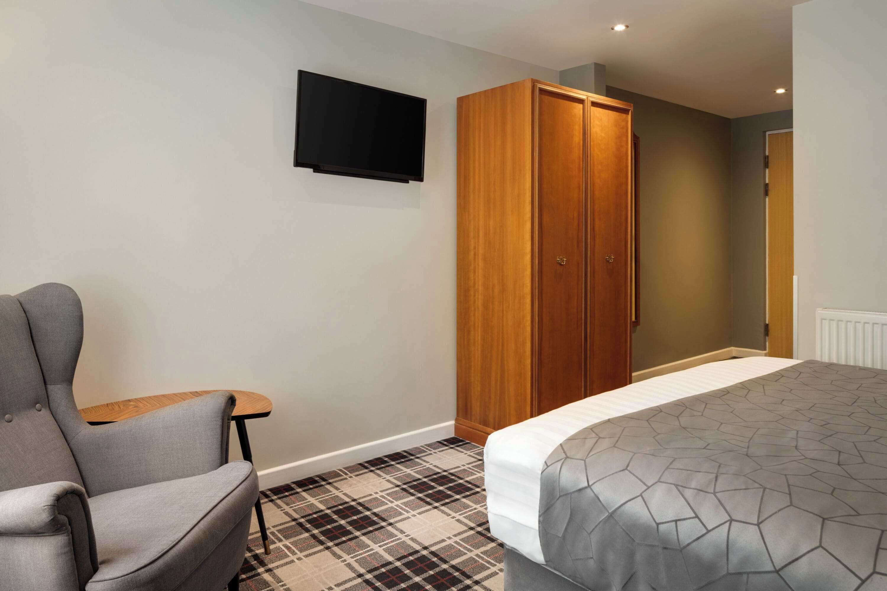 A hotel room with a bed, a grey armchair, a small table, a wardrobe, and a mounted television.