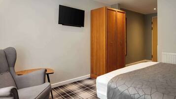 A hotel room with a bed, a grey armchair, a small table, a wardrobe, and a mounted television.