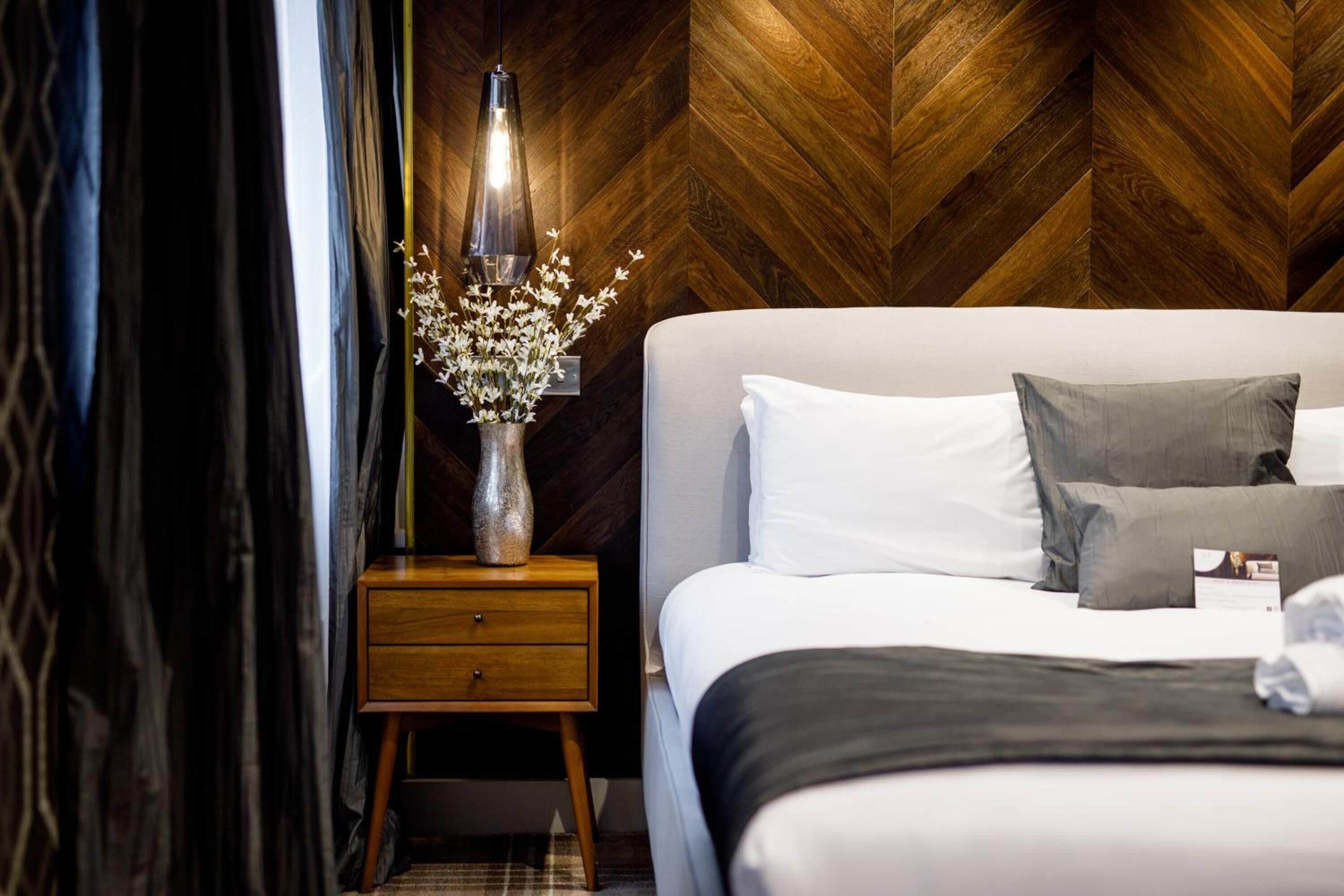 Un lit bien fait, avec une literie blanche et grise, une table de chevet en bois ornée d’un vase de fleurs et un mur décoré d’un motif en chevrons.