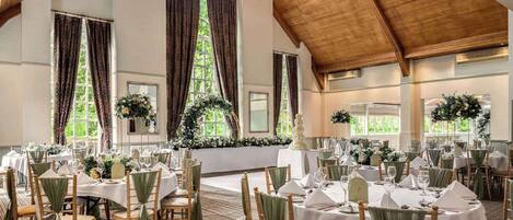 A banquet hall with multiple round tables set for dining, large windows with curtains, and a wooden ceiling.