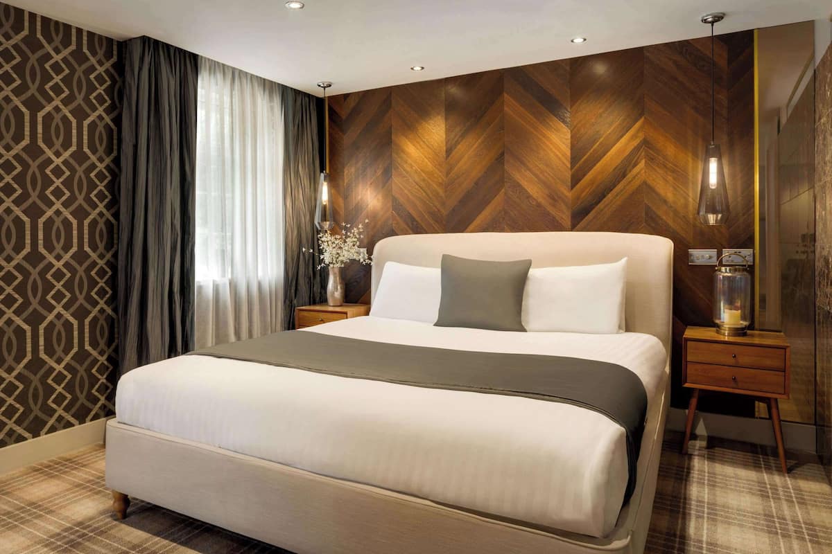 A modern hotel room with a large bed, a wooden herringbone-patterned wall, a nightstand with a lamp, and a decorative vase.