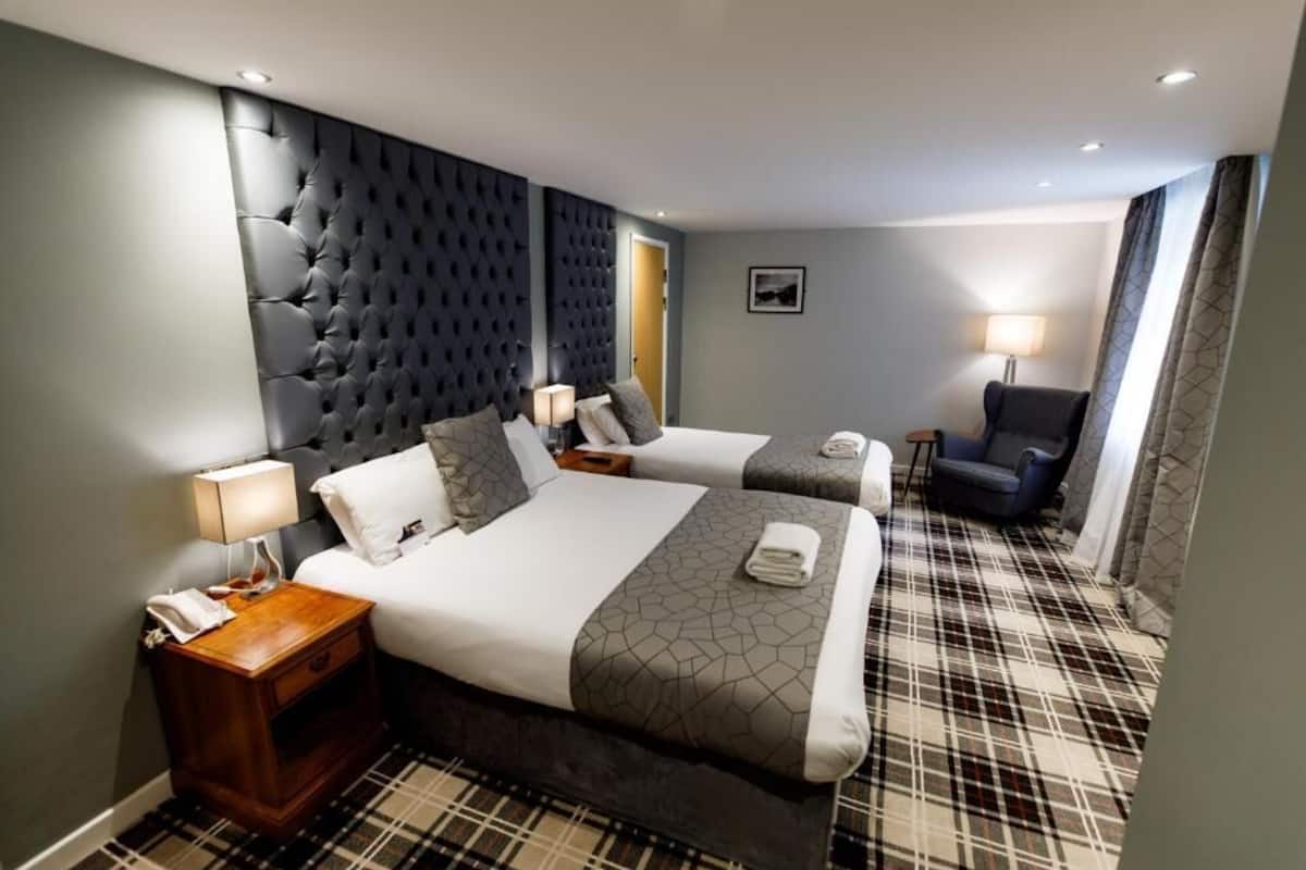 A hotel room with two beds, a dark tufted headboard, a plaid carpet, and a small seating area with a chair and table.