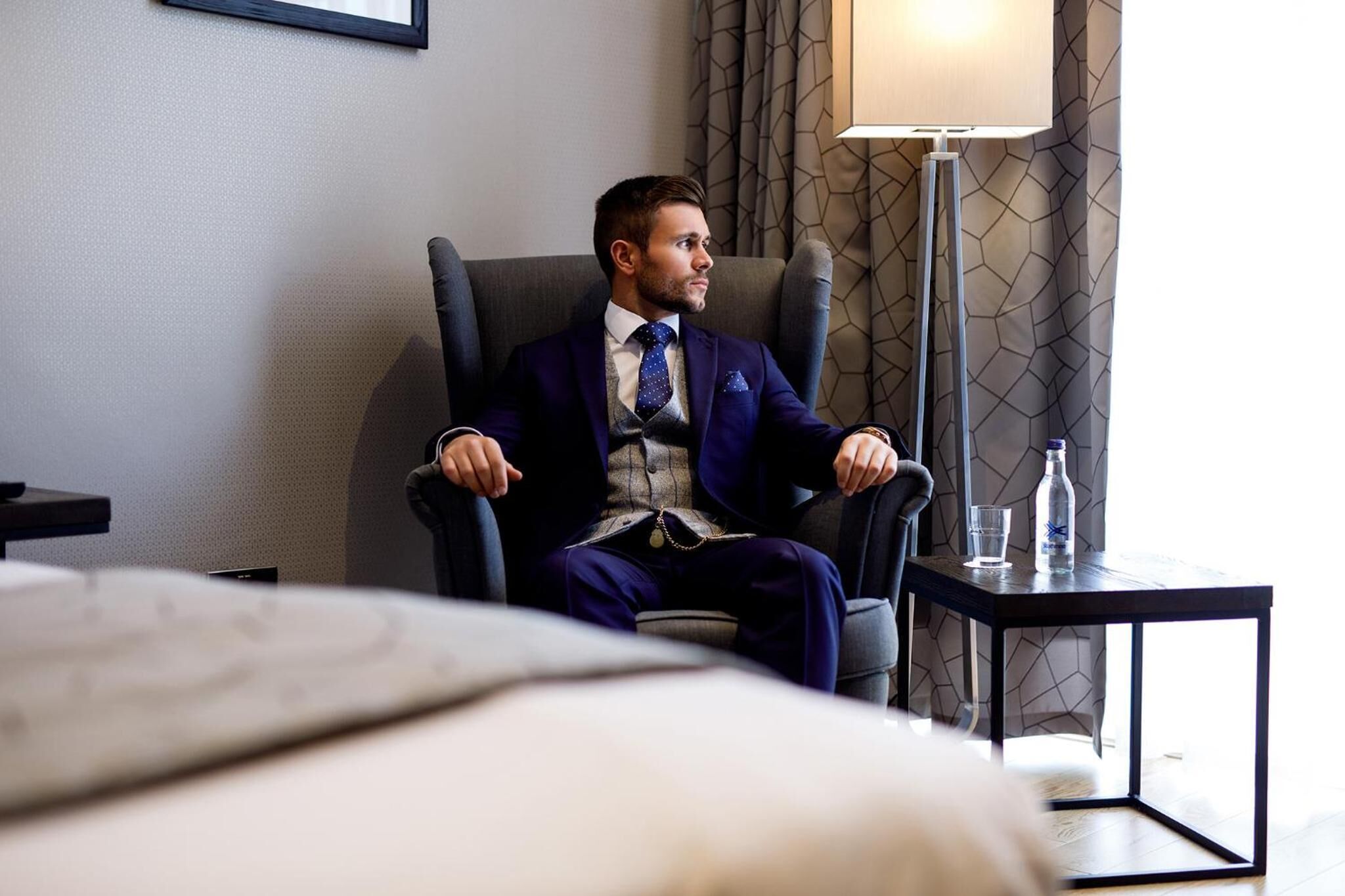 A man in a dark suit sitting in a modern armchair in a hotel room with a small table, a lamp, and a bottle of water.