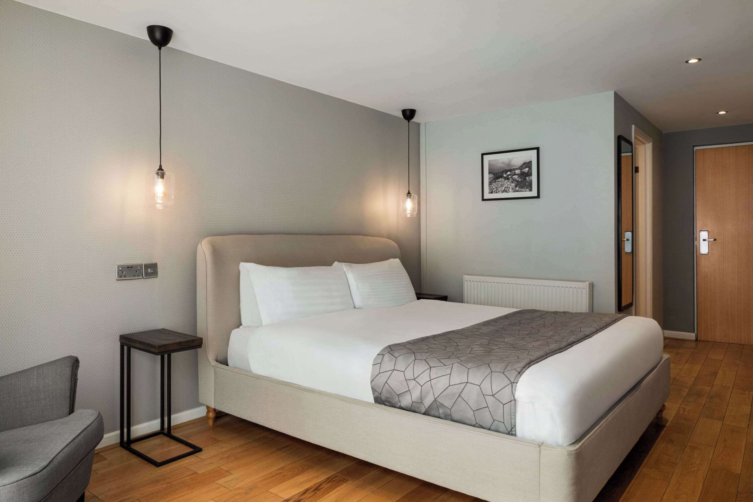 A modern bedroom with a large bed, a side table, a chair, and a hanging light fixture.