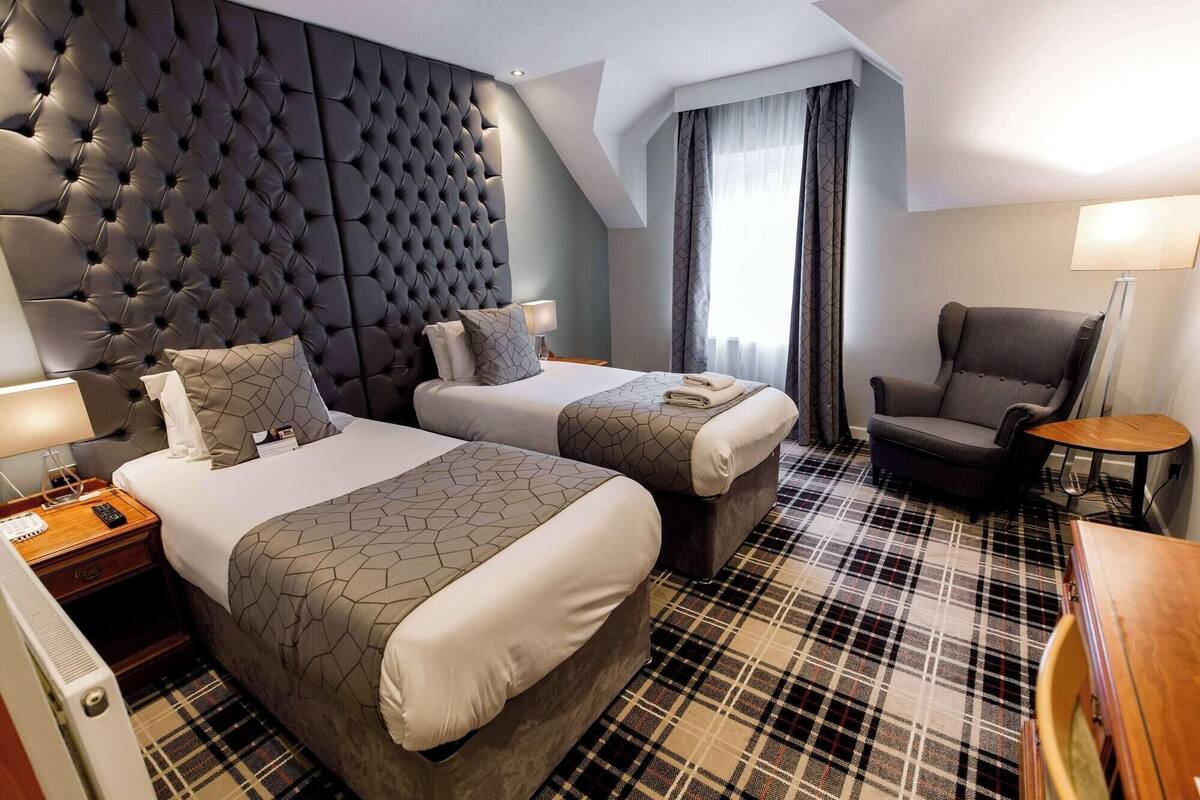 A hotel room with a tufted headboard, two beds with matching linens, a plaid carpet, a chair, and a small table.