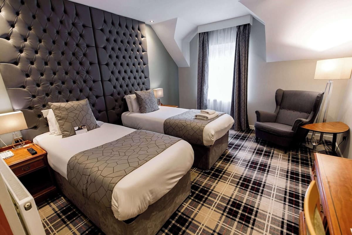 Habitación de hotel con cabecera capitonada, dos camas con ropa de cama a juego, alfombra escocesa, una silla y una pequeña mesa.