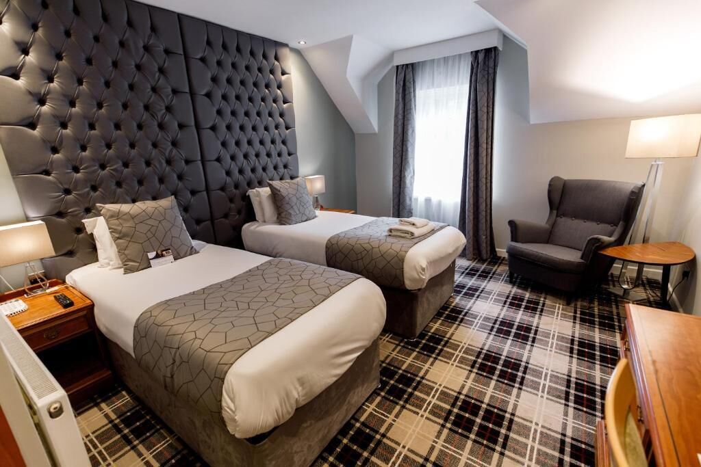 Una camera d'albergo con due letti, una testiera trapuntata, una moquette a quadri, una sedia e un tavolino.
