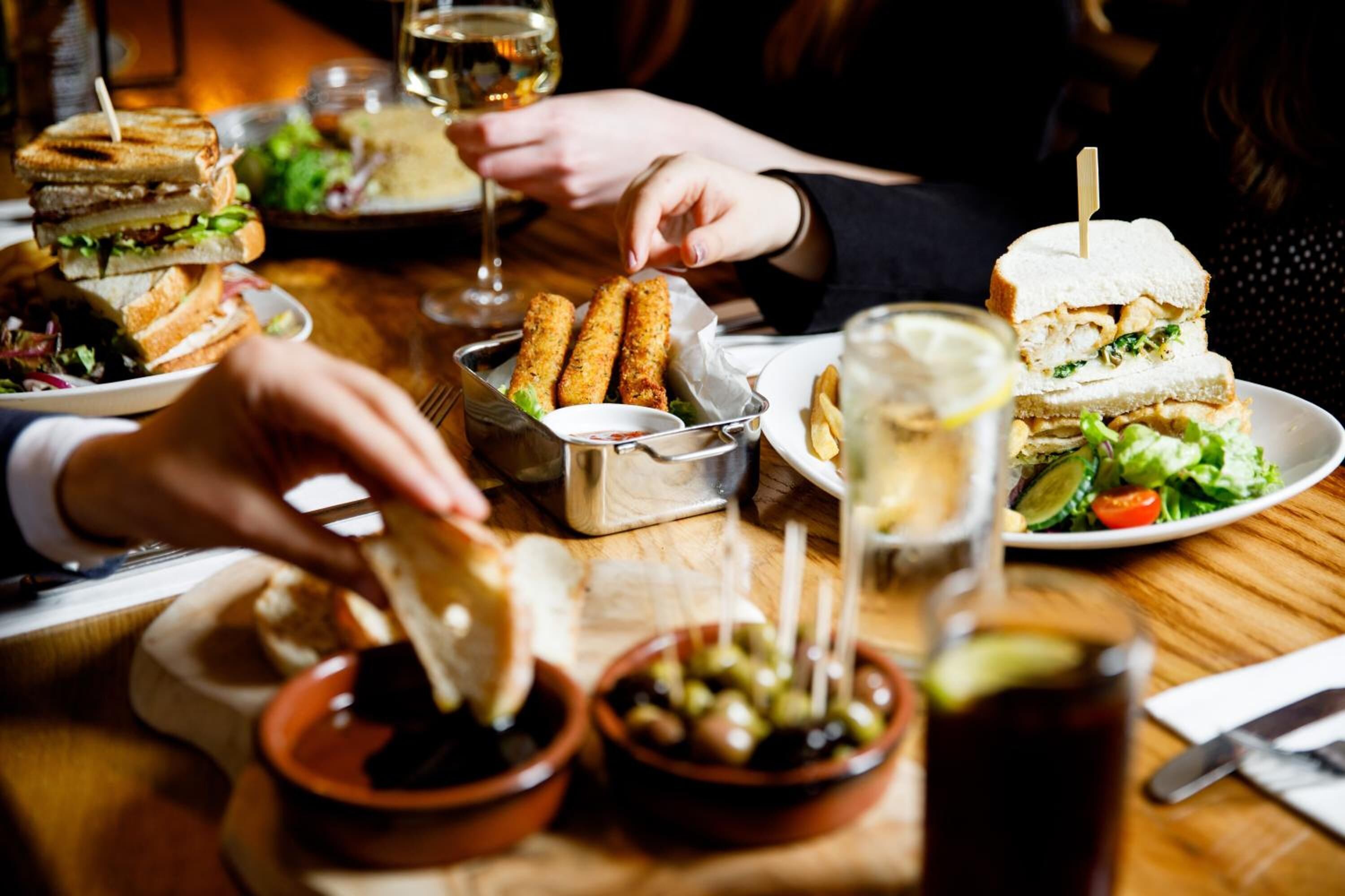 A dining table with sandwiches, a glass of wine, and small bowls of olives and sauces.