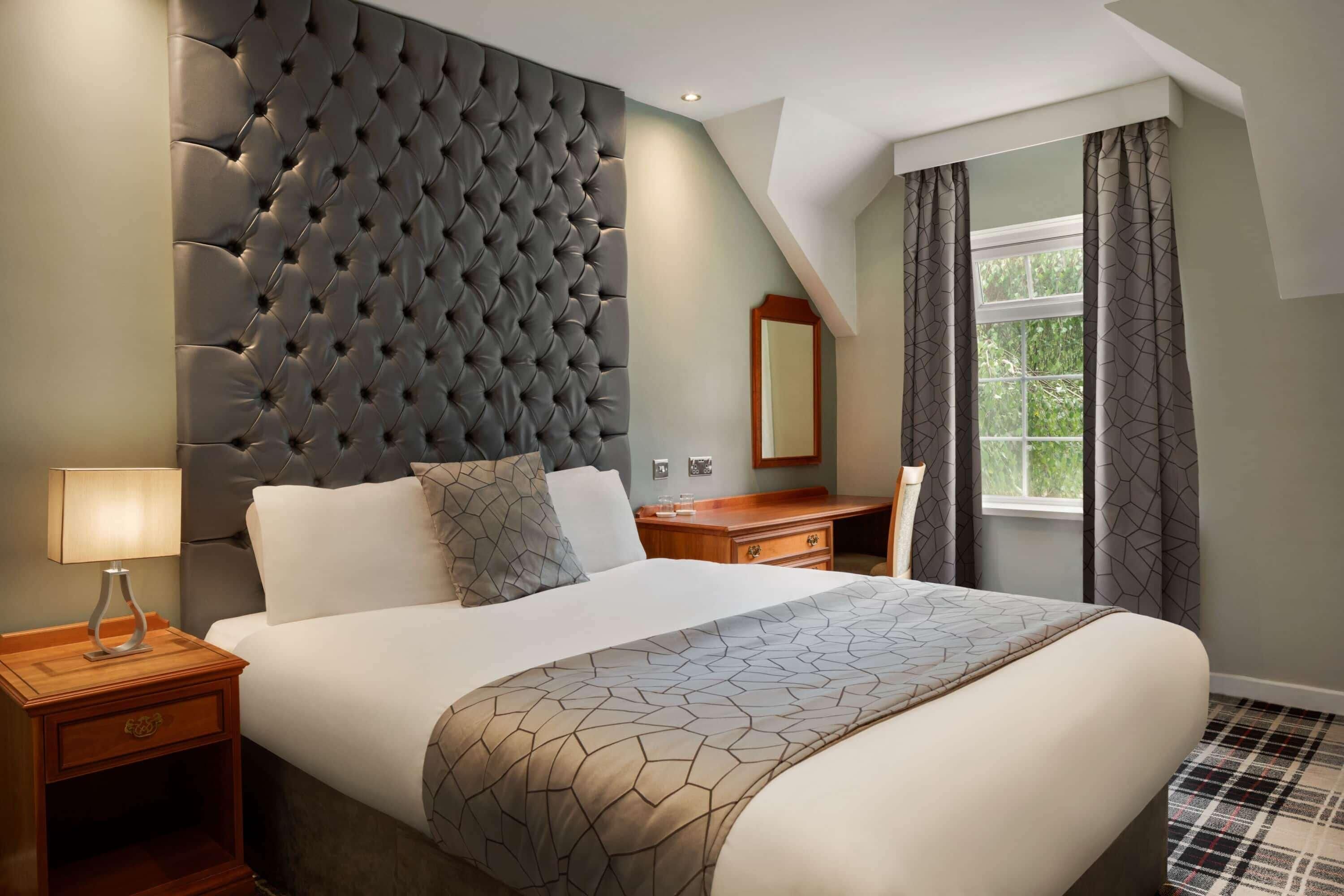 Une chambre à coucher avec une tête de lit capitonnée, un lit recouvert d’une couvre-lit à motifs géométriques, une table de chevet avec une lampe, une commode en bois et une fenêtre avec des rideaux.