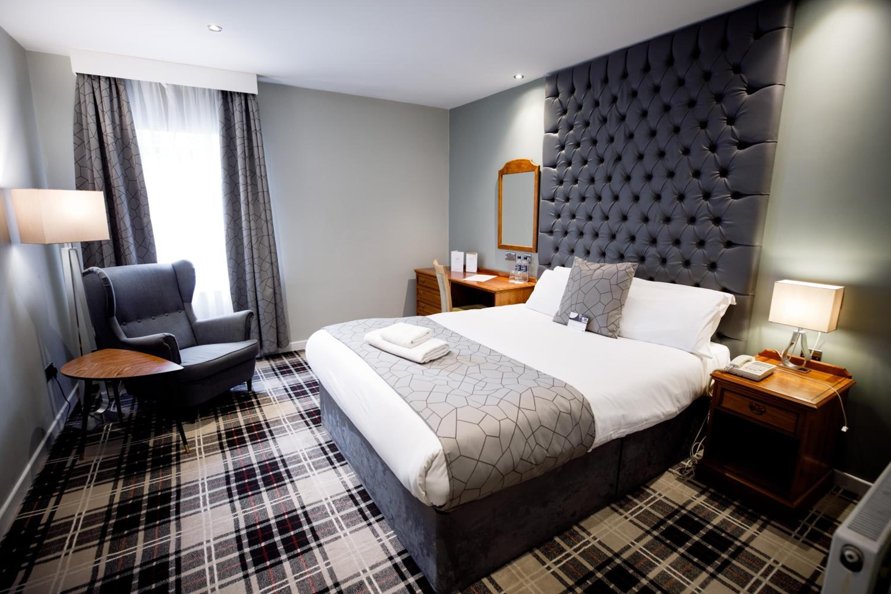 A hotel room with a large bed, a chair, a desk, a nightstand, and a window with curtains.