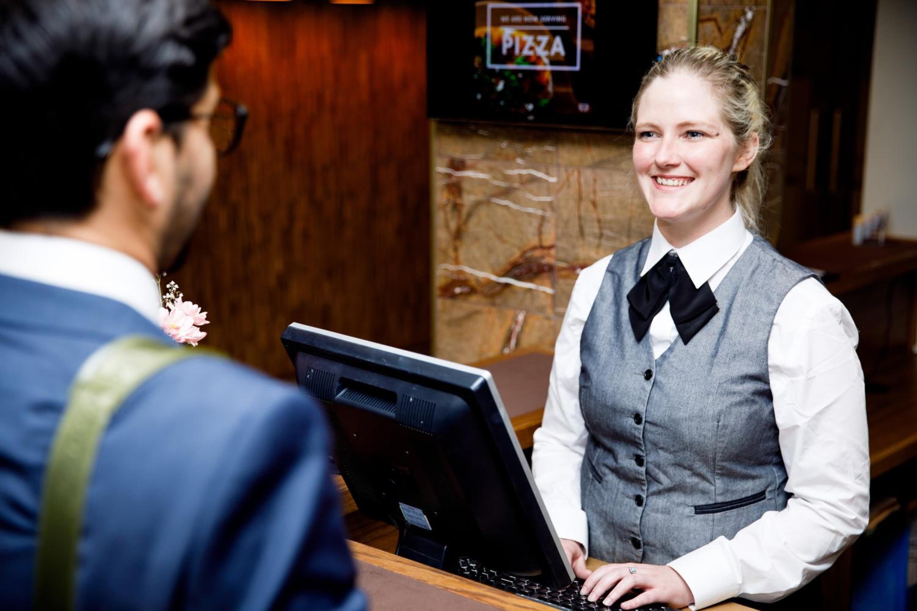 A person in a grey vest and white shirt with a black bow tie standing at a counter with a computer.