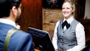 A person in a grey vest and white shirt with a black bow tie standing at a counter with a computer.