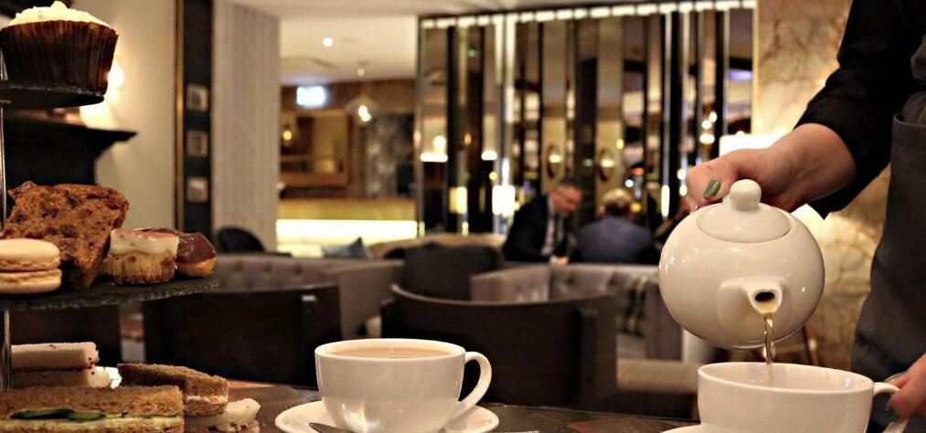 A person pouring tea into a cup, with pastries and a muffin on the table.