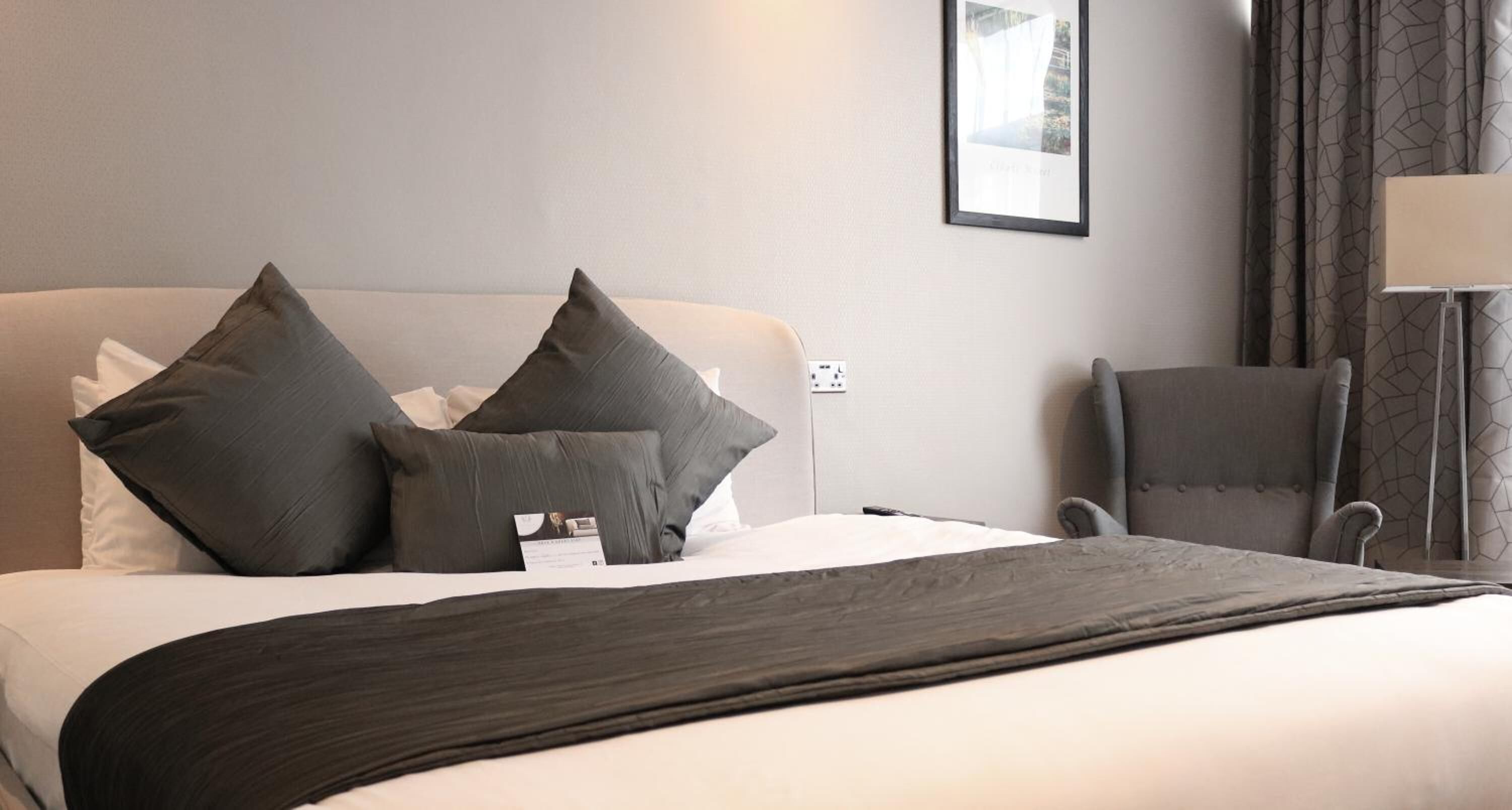 Habitación de hotel con cama, dos almohadas, una silla, una lámpara y un cuadro en la pared.