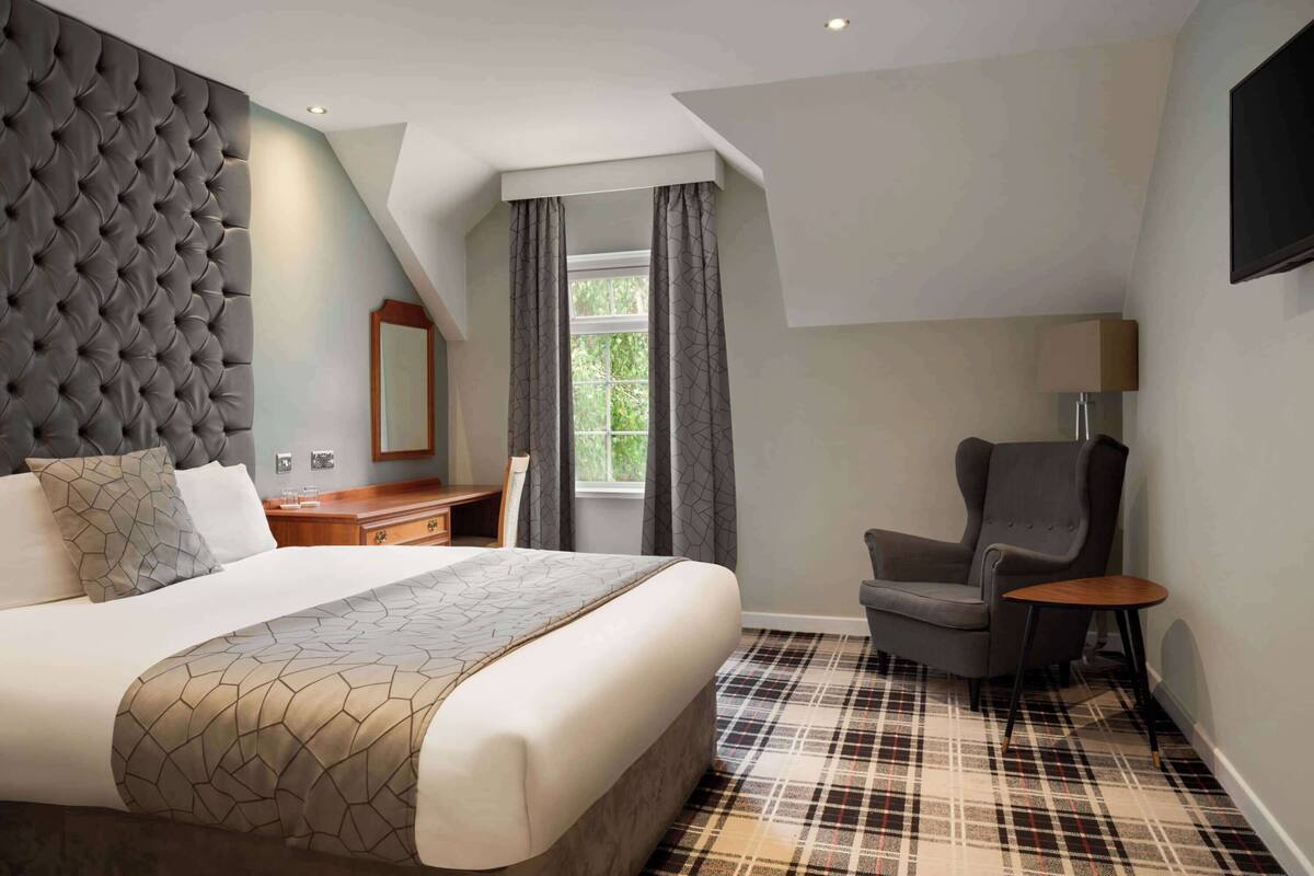 A hotel room with a large bed, a chair, a small table, a desk, and a mirror.