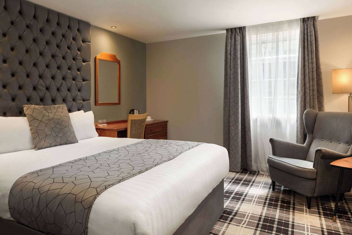A hotel room with a large bed, a tufted headboard, a desk with a chair, a lamp, and a view of the outdoors through a window with curtains.