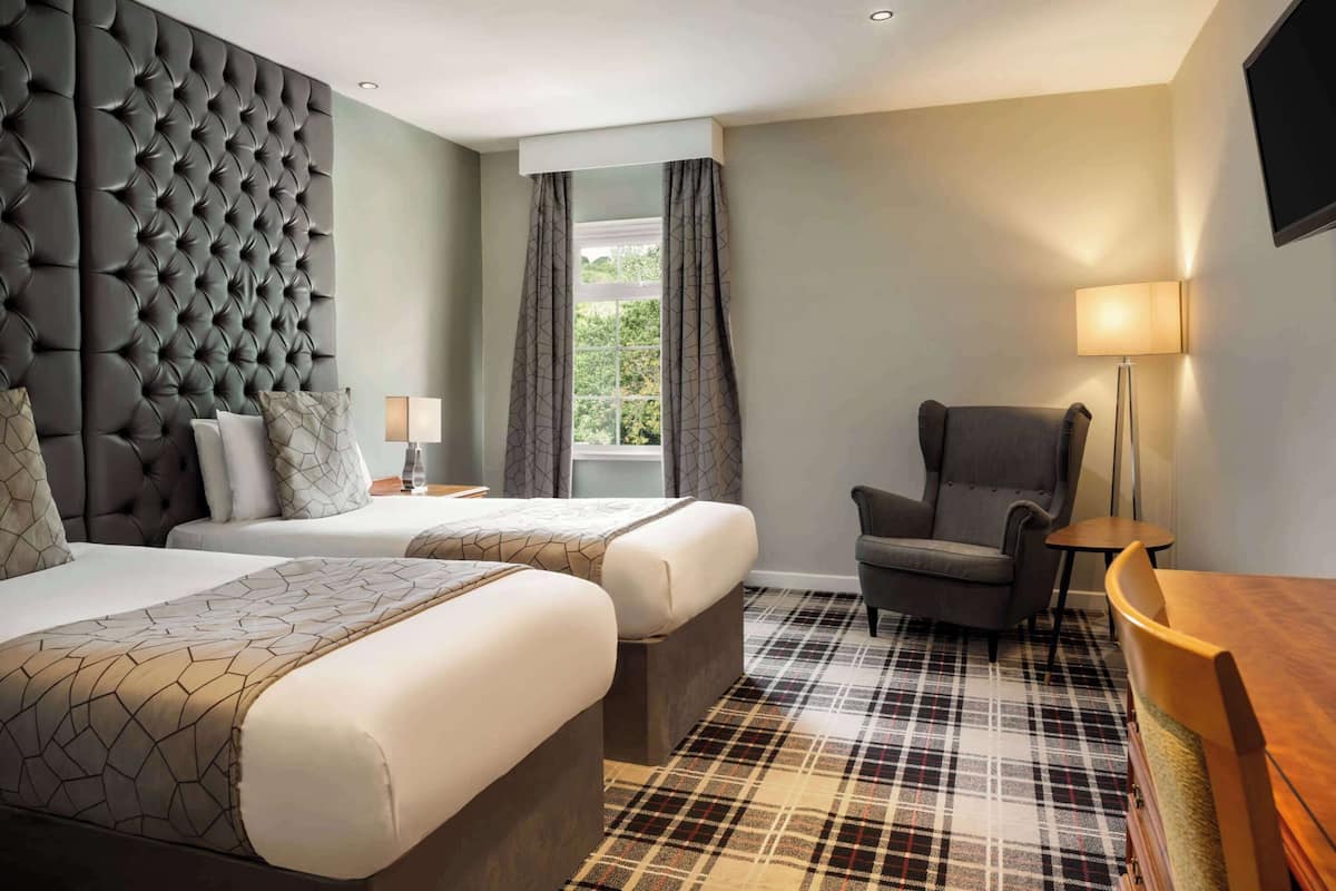 A hotel room with two beds, a large tufted headboard, a plaid carpet, a chair, a table, and a TV mounted on the wall.