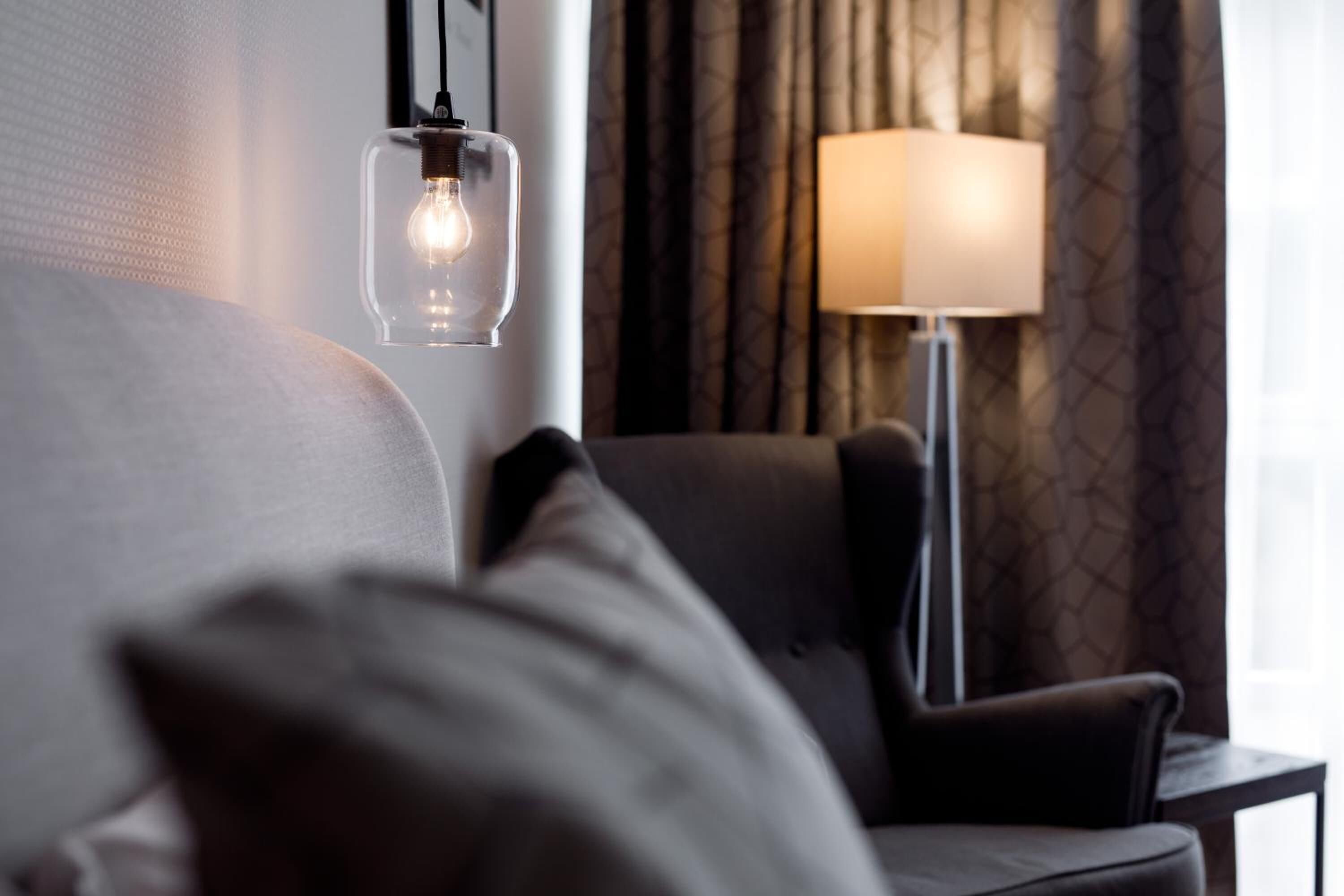 A modern hotel room with a clear light bulb hanging from the ceiling, a standing lamp, and a dark-colored sofa.