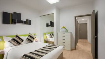 A modern hotel room with a bed, a nightstand, a dresser, a mirror, and a door leading to another room.