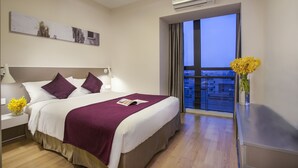 A hotel room with a large bed, a nightstand with a lamp, a window with a view, and an air conditioning unit. - Citadines Xinghai Suzhou (Suzhou)