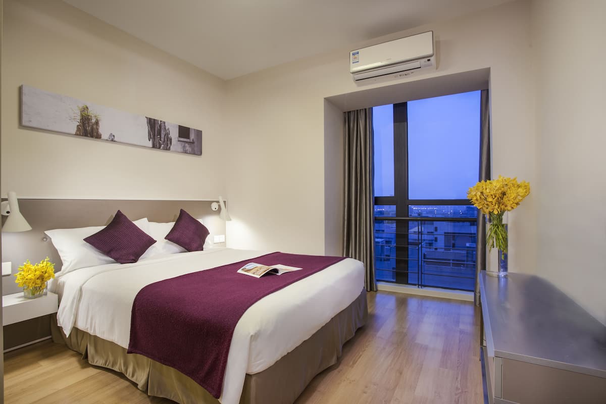 A hotel room with a large bed, a nightstand with a lamp, a window with a view, and an air conditioning unit.