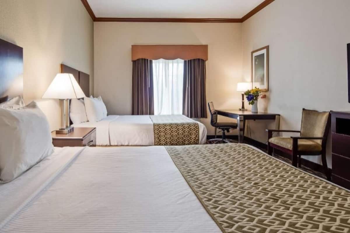 A hotel room with two beds, a desk, a chair, a lamp, and a window with curtains.