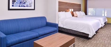Habitación de hotel con un sofá azul, una mesa de centro de madera, dos camas con ropa de cama blanca y una obra de arte abstracta enmarcada en la pared.