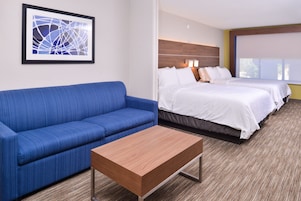 Habitación de hotel con un sofá azul, una mesa de centro de madera, dos camas con ropa de cama blanca y una obra de arte abstracta enmarcada en la pared.