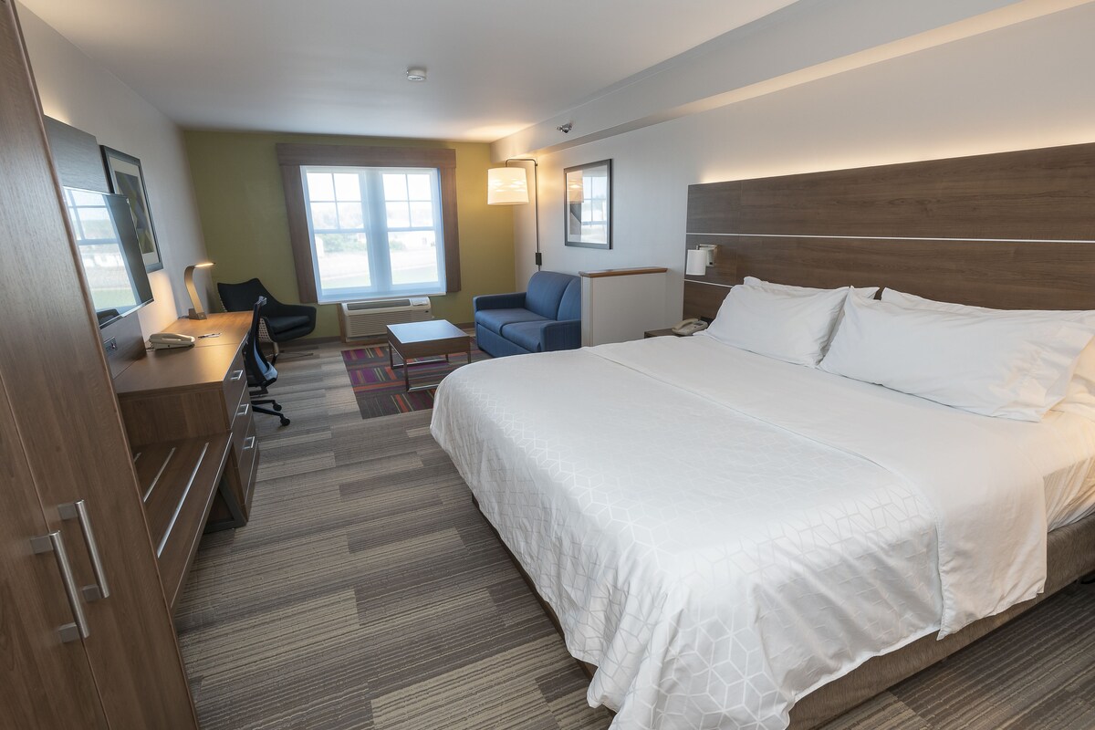 Ett hotellrum med en stor säng, ett skrivbord med en stol, ett litet bord och ett fönster med gardiner.