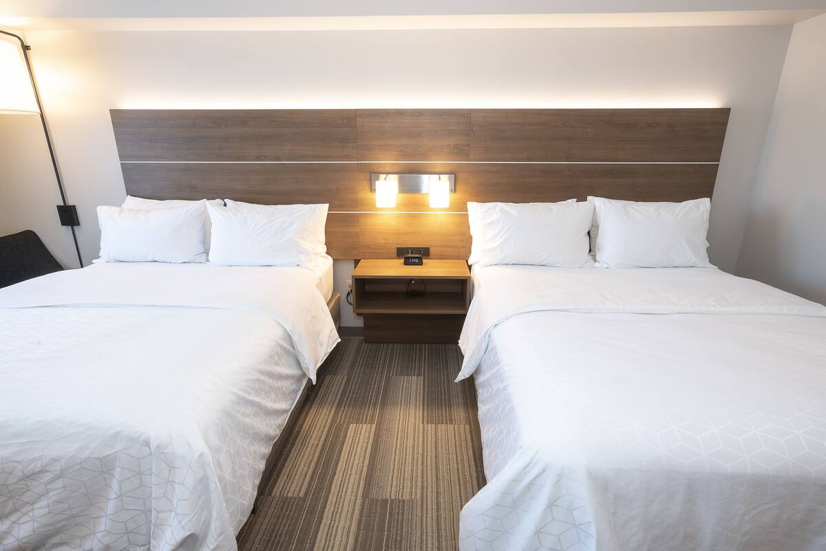 Habitación de hotel con dos camas, cabecero de madera, lámparas de noche y mesita de noche con un reloj.