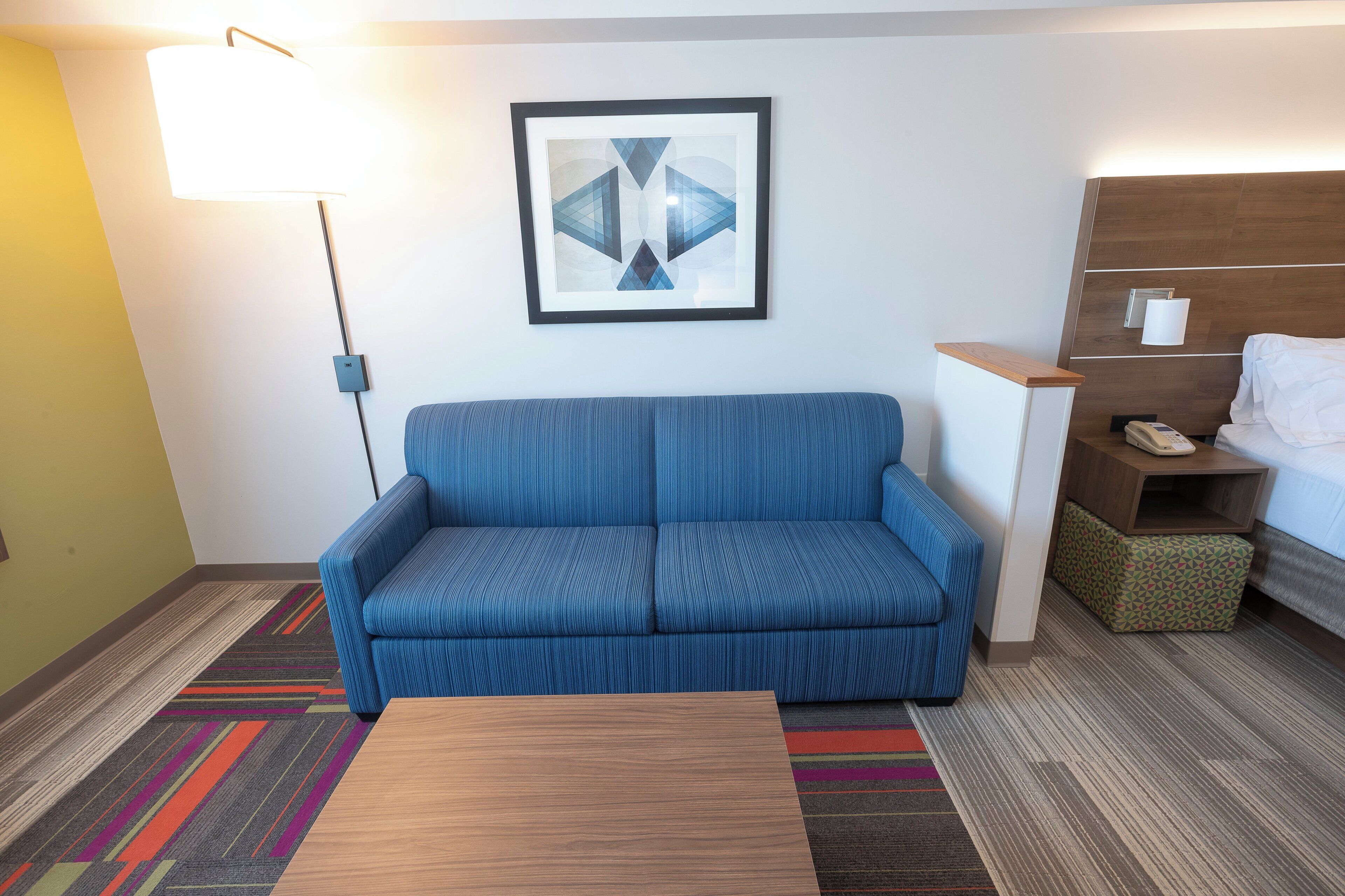 A hotel room with a blue sofa, a wooden coffee table, a framed abstract artwork on the wall, a bedside table with a lamp, and a bed with a patterned pillow.