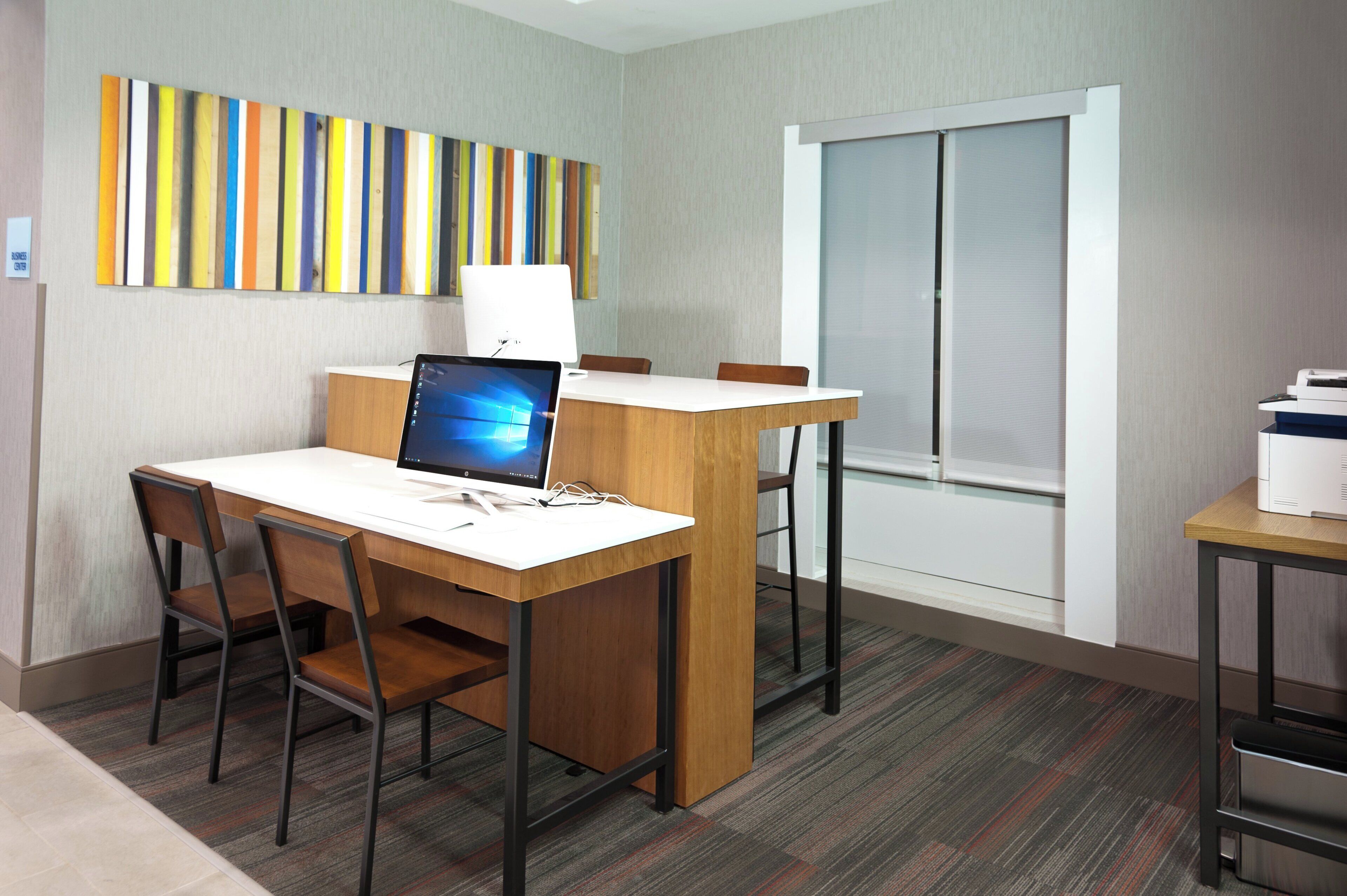 Habitación de hotel con escritorio, silla y computadora. Se aprecia una ventana con persianas, un cuadro con motivos rayados en la pared y piso alfombrado.