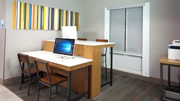 Habitación de hotel con escritorio, silla y computadora. Se aprecia una ventana con persianas, un cuadro con motivos rayados en la pared y piso alfombrado.