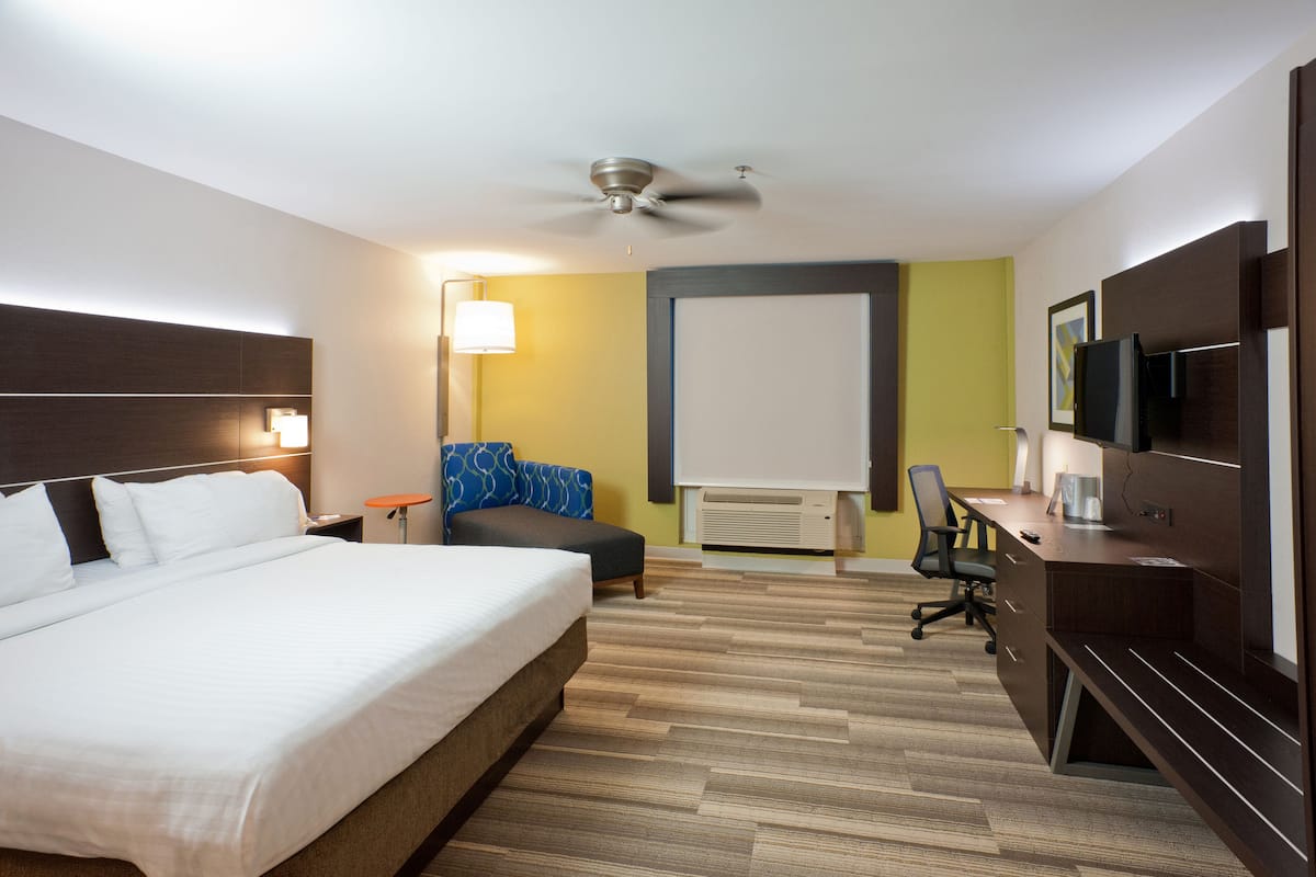 A hotel room with a large bed, a desk with a chair, a ceiling fan, a TV, and a lamp.
