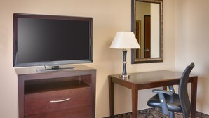 Quarto de hotel com televisão de tela plana, escrivaninha com luminária, cadeira e espelho na parede.