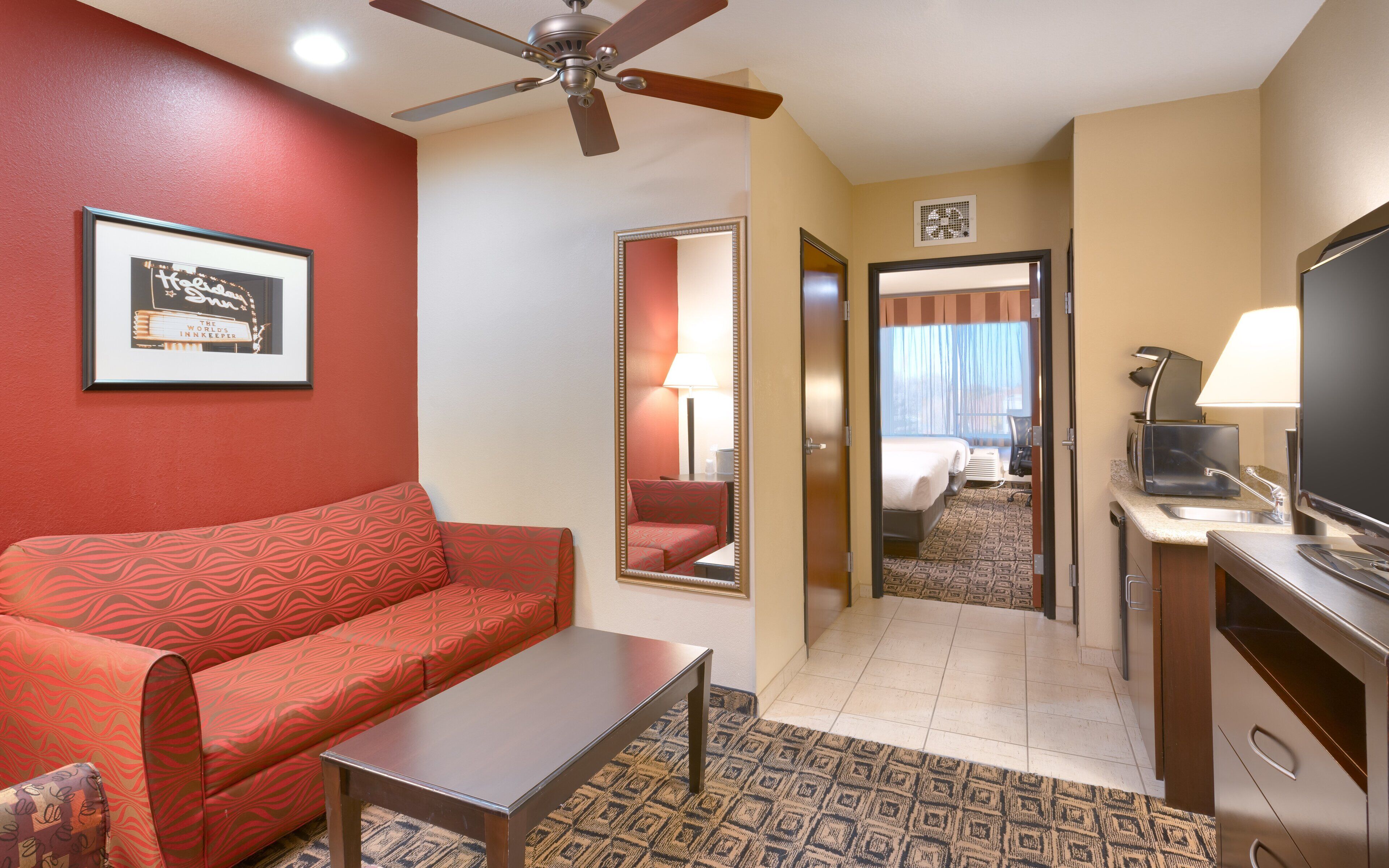 A hotel room with a red sofa, a small table, a ceiling fan, a television, and a mirror reflecting the room.