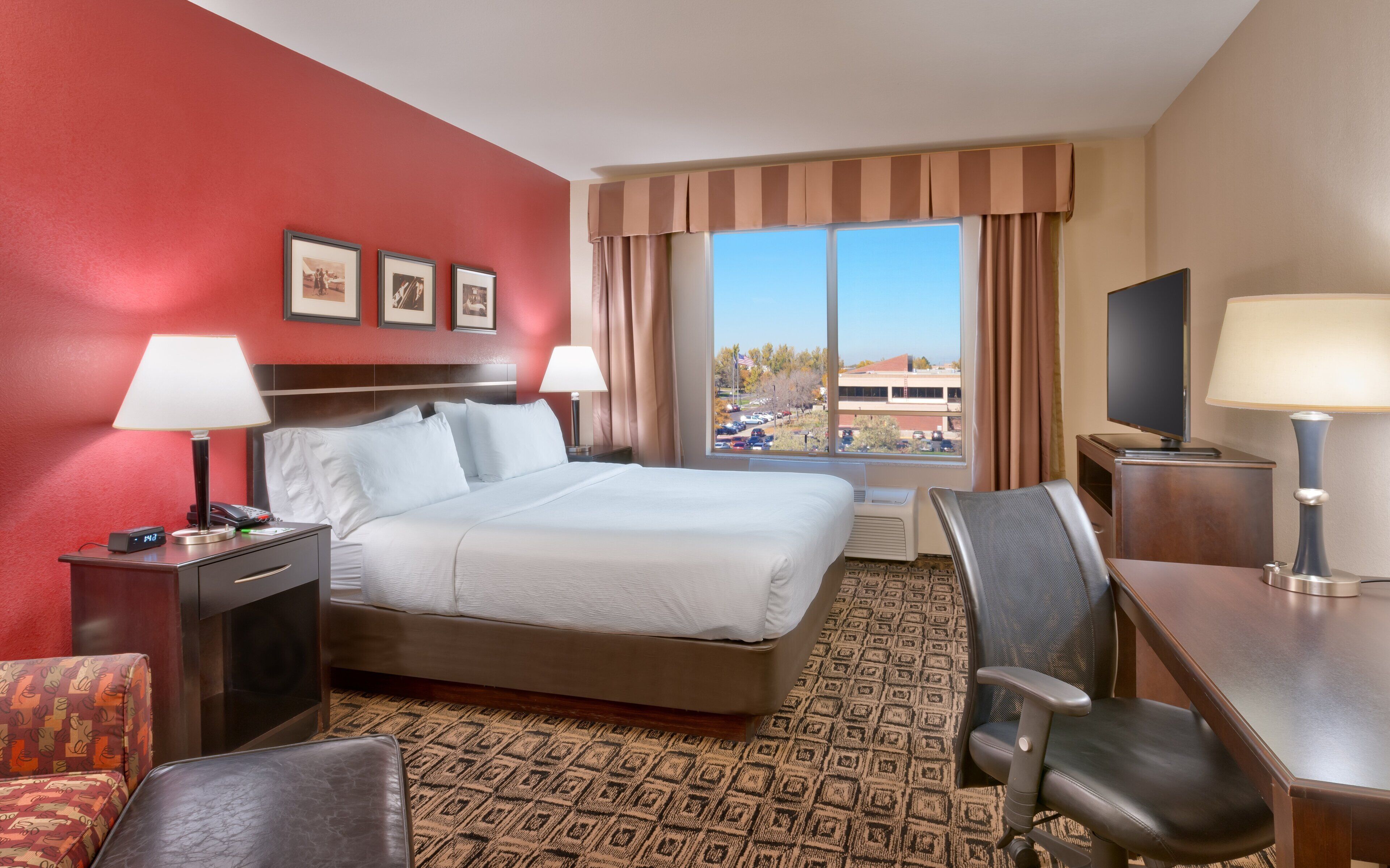 Ein Hotelzimmer mit Bett, Schreibtisch, Stuhl und einem großen Fenster mit Blick auf einen Parkplatz und Gebäude.